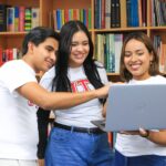 Tres jóvenes consultan una computadora portátil en una biblioteca.