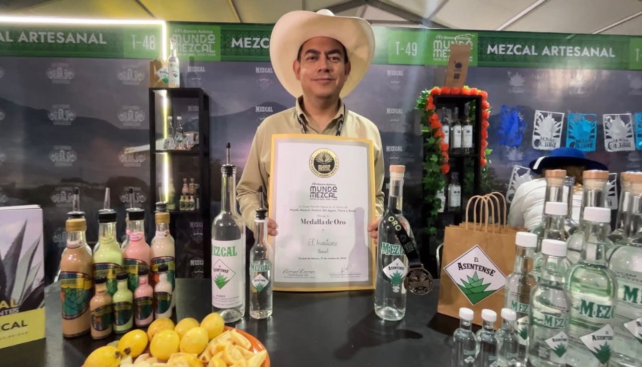 Representante de una marca de mezcal muestra un reconocimiento en el festival Mundo Mezcal 2026, junto a botellas de mezcal artesanal.