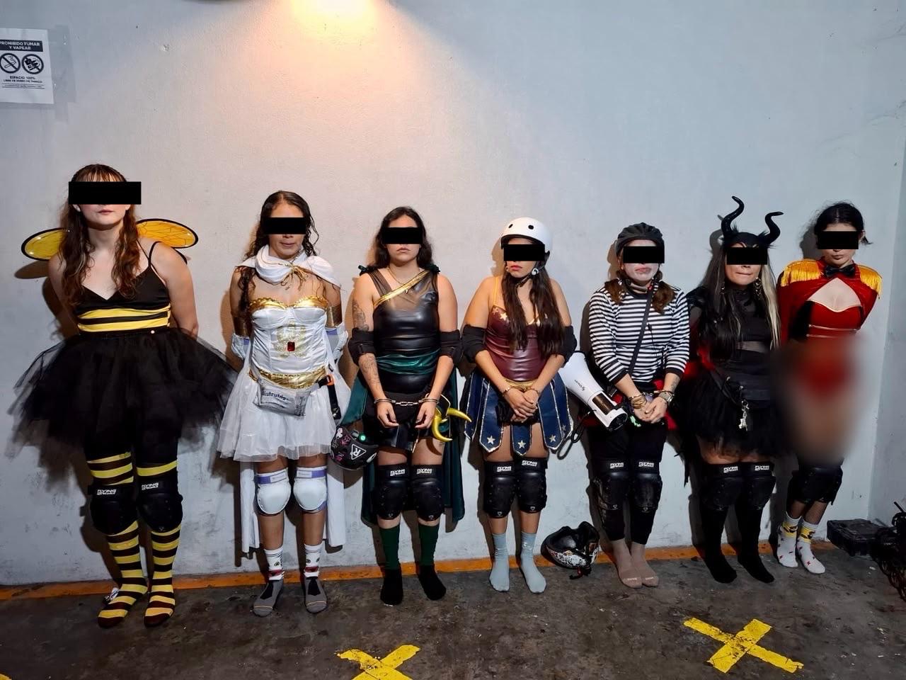 Siete personas con disfraces y patines aparecen de pie ante una pared; sus ojos están cubiertos por franjas negras.