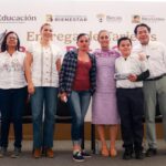 Autoridades educativas y asistentes posan durante una entrega de tarjetas de la Beca Rita Cetina, ante un fondo de programas de becas.