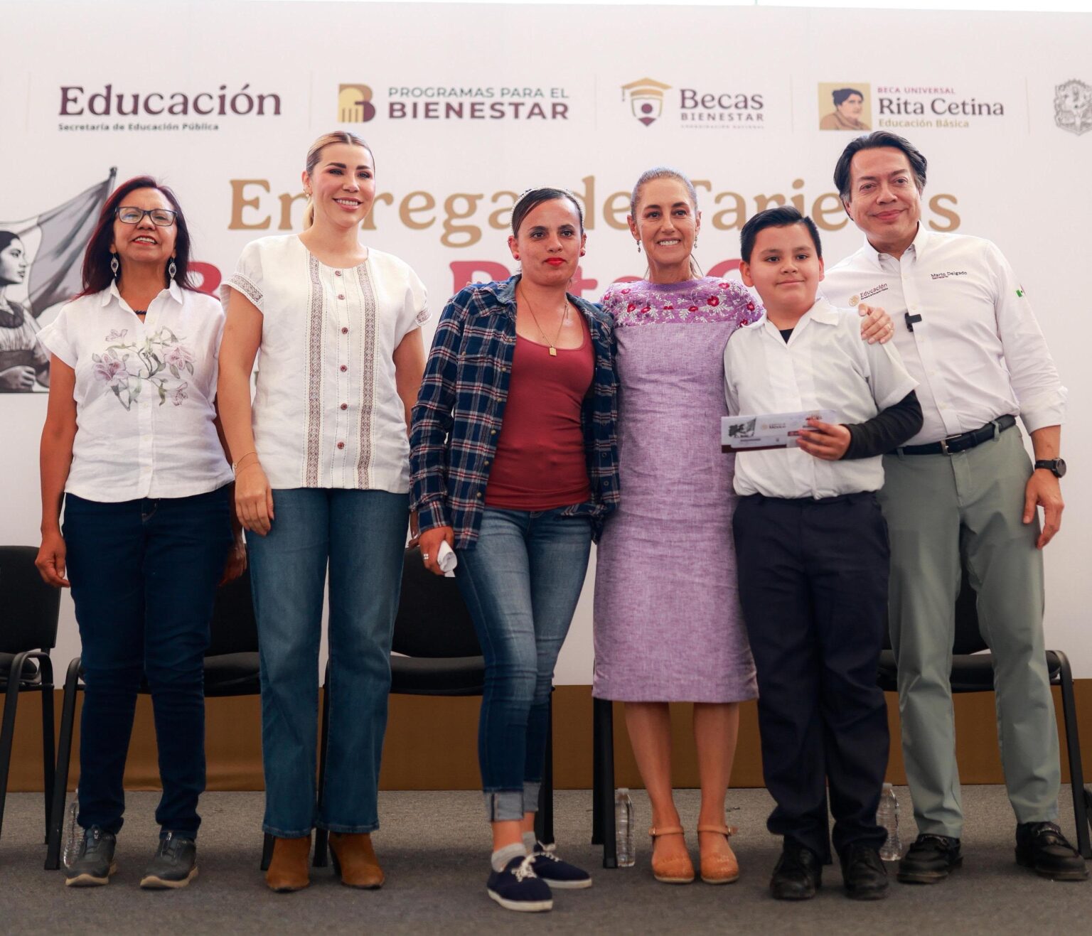 Autoridades educativas y asistentes posan durante una entrega de tarjetas de la Beca Rita Cetina, ante un fondo de programas de becas.