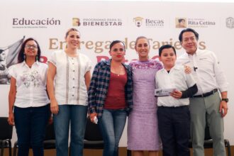 Autoridades educativas y asistentes posan durante una entrega de tarjetas de la Beca Rita Cetina, ante un fondo de programas de becas.