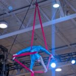 Atleta realiza una figura acrobática suspendida en telas aéreas rosas durante una competencia en un recinto deportivo.