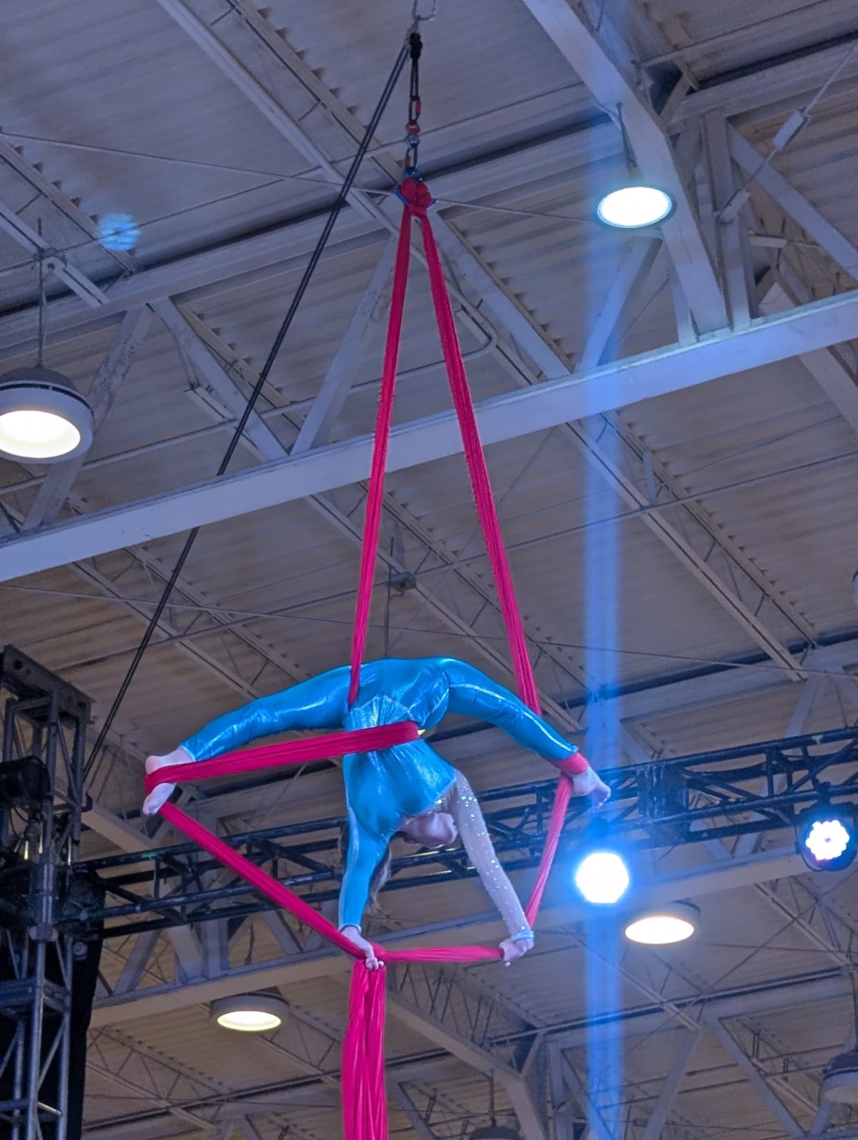 Atleta realiza una figura acrobática suspendida en telas aéreas rosas durante una competencia en un recinto deportivo.