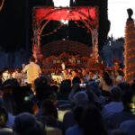 Concierto ante un altar de Día de Muertos decorado con flores de cempasúchil en un panteón, con público reunido al anochecer.