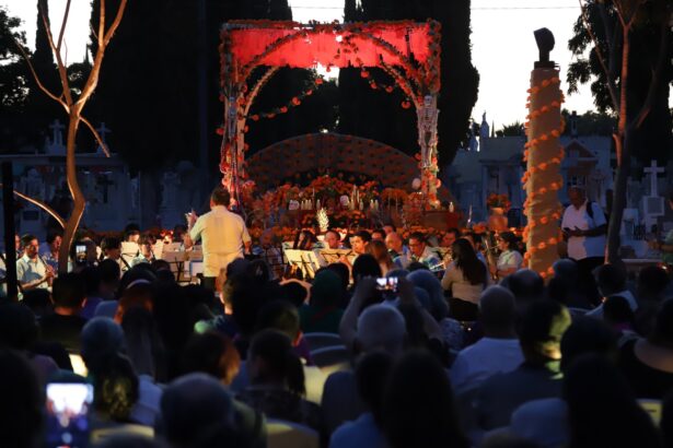 Concierto ante un altar de Día de Muertos decorado con flores de cempasúchil en un panteón, con público reunido al anochecer.