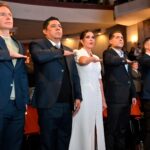 Asistentes a un acto institucional en un teatro, de pie y con el brazo derecho extendido durante una ceremonia.