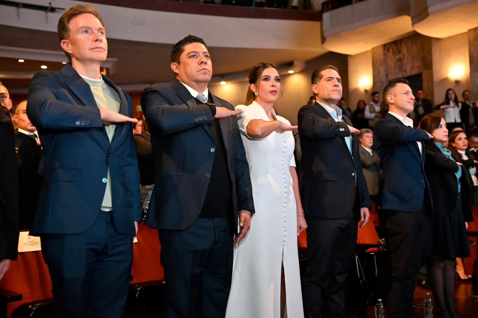Asistentes a un acto institucional en un teatro, de pie y con el brazo derecho extendido durante una ceremonia.
