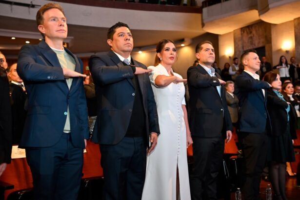 Asistentes a un acto institucional en un teatro, de pie y con el brazo derecho extendido durante una ceremonia.