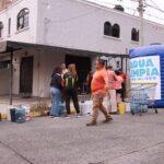 Habitantes se reúnen con recipientes para recibir agua junto a una cisterna azul con el letrero Agua Limpia para Jalisco en una calle de Guadalajara